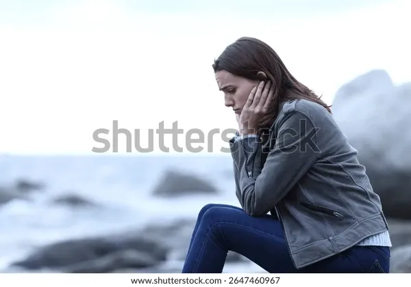 Eine Frau sitzt nachdenklich auf Felsen am Meer und stützt den Kopf in die Hand