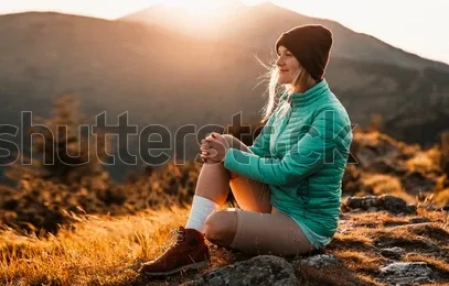 Eine Frau in grüner Jacke sitzt bei Sonnenuntergang auf einer Bergwiese